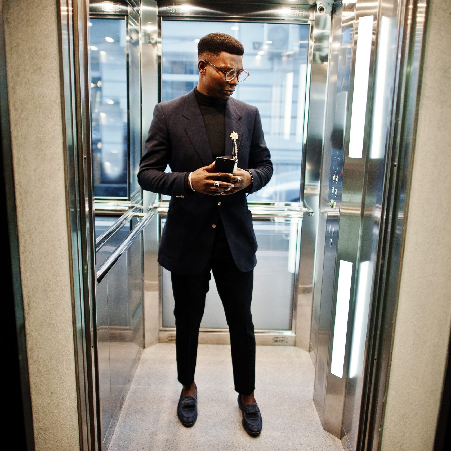 Fashionable african american man in suit and glasses with mobile phone and cup of coffee at hands posed inside elevator.